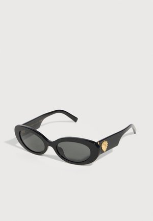 Μαύρα οβάλ γυαλιά ηλίου με σκουρόχρωμους φακούς και χρυσό έμβλημα λιονταριού στον βραχίονα, εμφανίζονται σε λευκό φόντο.