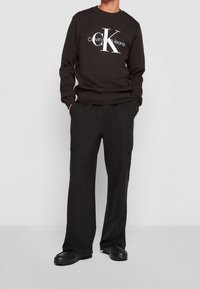 Černá mikina s logem Calvin Klein Jeans v bílé barvě, doplněná černými širokými kalhotami typu cargo s bočními kapsami. Hladký materiál a volný střih.