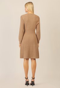 Beige Strickkleid mit langen Ärmeln und einem figurbetonten Oberteil, das mit einem Zopfmuster verziert ist und zu einem knielangen Rock ausläuft.