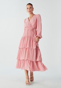 Dlouhé, růžové šaty s volánky vyrobené z lehkého materiálu, s překříženým výstřihem do V, dlouhými rukávy a nastavitelným páskem v pase.