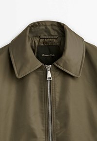 Veste vert olive avec col pointu, fermeture éclair argentée visible et étiquette noire portant l'inscription "Massimo Dutti" à l'intérieur du col.