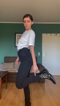 Calças de ganga pretas justas com cintura alta, combinadas com uma t-shirt branca. Botins pretos com fivelas metálicas. Fundo interior simples.