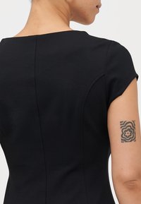 Svart tettsittende kjole med korte ermer, som har en glatt tekstur og rene linjer. Lite blomster-tatovering synlig på overarmen.
