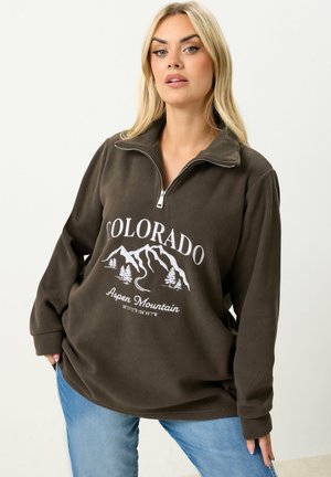 Frau mit blondem Haar, die ein dunkelgraues Fleecepullover mit halbem Reißverschluss trägt, auf dem "Colorado Aspen Mountain" und eine Berggrafik abgebildet sind, kombiniert mit blauen Jeans.
