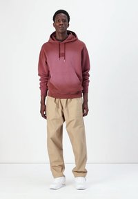 Maroon hoodie with kangaroo pocket, beige pants with pockets, and white sneakers. Simple design and relaxed fit with visible stitching details.