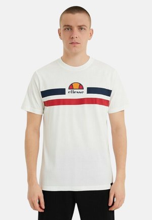 Jeune homme portant un t-shirt blanc avec des rayures horizontales bleu marine et rouges et un logo ellesse sur la poitrine, debout devant un fond uni.