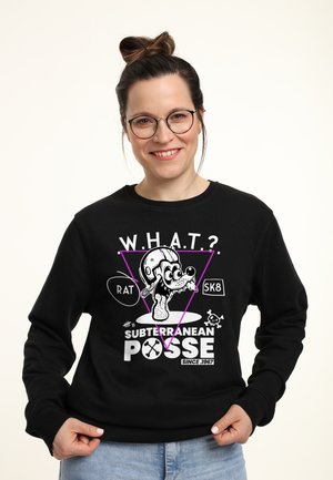 Uśmiechnięta kobieta w okularach i czarnej bluzie z nadrukiem kreskówkowego psa w hełmie oraz napisem "W.H.A.T.? Subterranean Posse Since 1967."