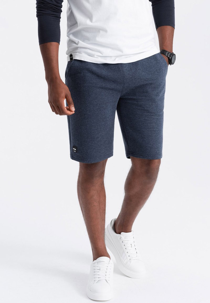 Pantaloni corti blu navy realizzati in materiale texturizzato, dotati di una tasca laterale e di una piccola etichetta con logo. Abbinati a sneakers bianche.