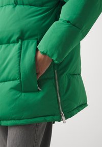 Grüne Pufferjacke mit gestepptem Design, ausgestattet mit einer Seitentasche und einem silbernen Reißverschluss. Weicher Stoff, mittellang und mit lockerer Passform.