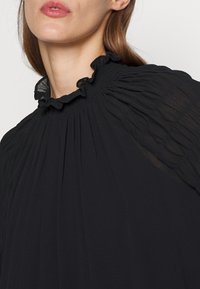Blusa nera plissettata con un collo alto e arricciato. Tessuto leggero con una sottile trasparenza e dettagli di texture delicati.
