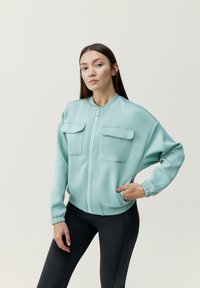 Jasno miętowa kurtka bomber z dwiema kieszeniami na piersi, ściągaczami na rękawach i zamek błyskawiczny z przodu, zestawiona z czarnymi legginsami. Gładki, elastyczny materiał.