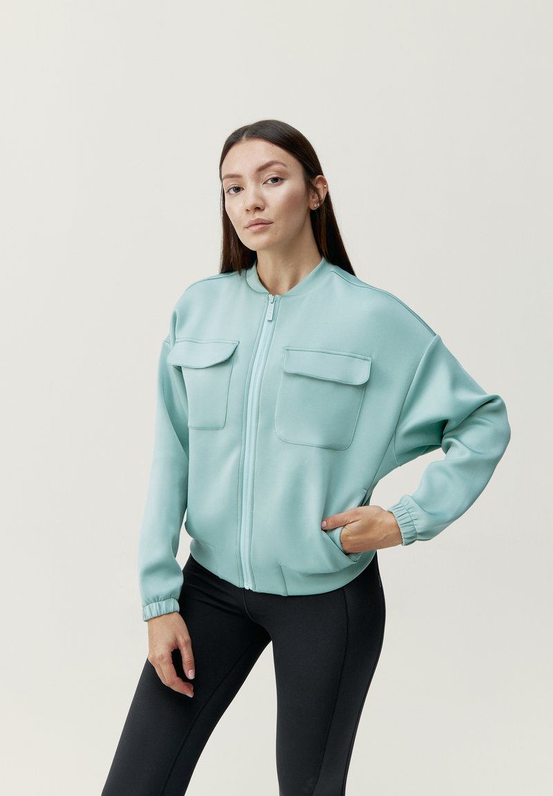 Jasno miętowa kurtka bomber z dwiema kieszeniami na piersi, ściągaczami na rękawach i zamek błyskawiczny z przodu, zestawiona z czarnymi legginsami. Gładki, elastyczny materiał.