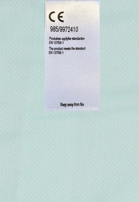 Ljusblått tyg med en texturerad yta. En vit etikett har CE-märkningen, produktnumret och en brandsäkerhetsvarning tryckt i svart.