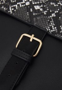 Schwarze Lederhandtasche mit einem Klappdeckel im Schlangenledermuster, ausgestattet mit einer goldenen metallischen Schnalle und einer strukturierten Oberfläche. Der Trageriemen hat verstellbare Löcher.