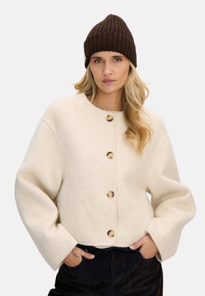 Kremowa wełniana kurtka z okrągłym dekoltem, oversize'owymi rękawami i dużymi drewnianymi guzikami, zestawiona z ciemnobrązową prążkowaną czapką beanie.