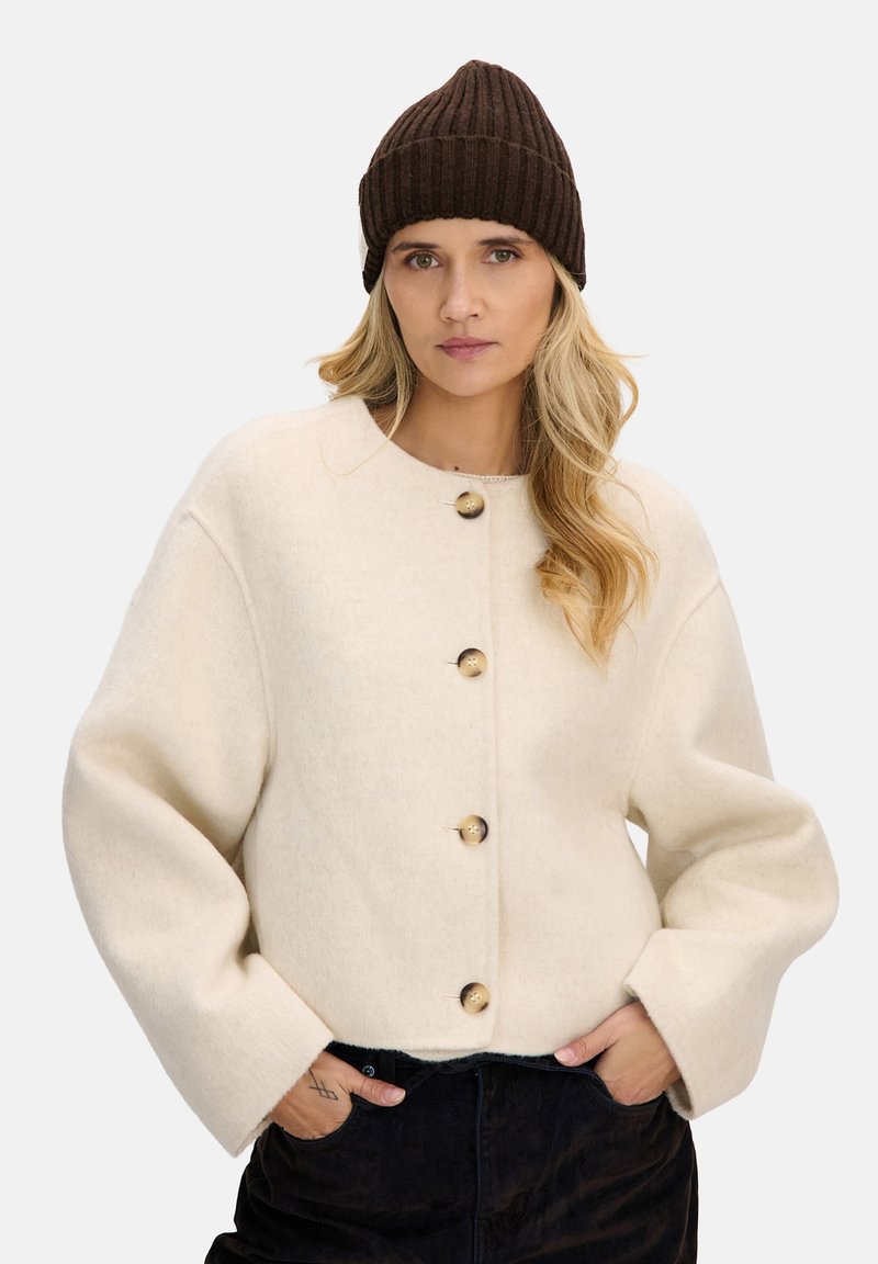 Kremowa wełniana kurtka z okrągłym dekoltem, oversize'owymi rękawami i dużymi drewnianymi guzikami, zestawiona z ciemnobrązową prążkowaną czapką beanie.