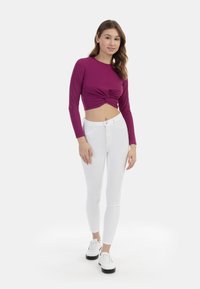 Tætsiddende, langærmet, lilla crop top med et snoet frontdesign, parret med hvide skinny jeans og hvid sneakers med sorte såler.