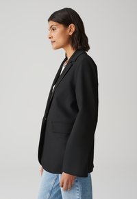 Zwarte blazer met een gestructureerd ontwerp, eenknopsluiting en twee voorzakken. Gecombineerd met lichtblauwe, hoge taille jeans.