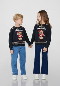 Zwei Kinder halten Händchen und tragen schwarze Weihnachtspullover mit einem Teddy-Bären-Motiv, blaue Jeans und weiße Schuhe.