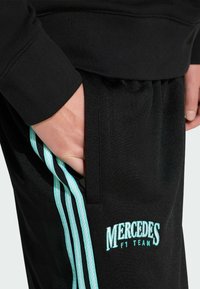 Schwarze Sporthose mit hellblauen Akzenten, mit einem Logo, seitlichem Streifendesign und einer Vordertasche mit sichtbarer Hand.