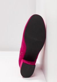 Chaussure en daim rose vif avec un petit talon carré, vue par le dessous avec une semelle noire texturée sur fond blanc.