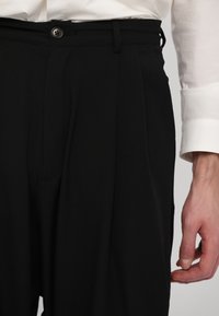 Pantaloni neri su misura con vestibilità ampia, dotati di pieghe frontali, chiusura con bottone e tasche, abbinati a una camicia bianca con il polsino visibile.