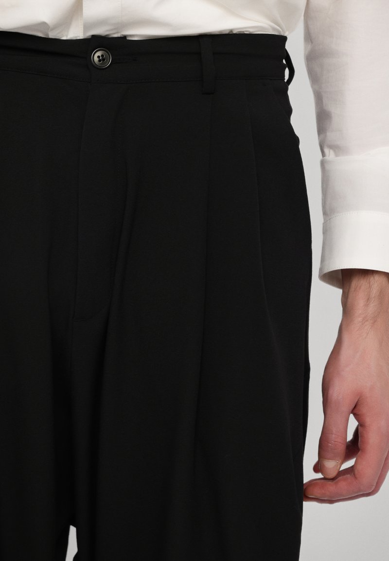 Pantaloni neri su misura con vestibilità ampia, dotati di pieghe frontali, chiusura con bottone e tasche, abbinati a una camicia bianca con il polsino visibile.