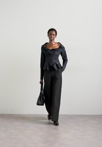 Sort skræddersyet bluse med off-shoulder design og knapdetaljer, parret med brede sorte bukser. Modellen holder en lille sort håndtaske.
