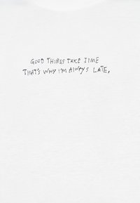 Λευκό ύφασμα με μαύρο χειρόγραφο κείμενο που αναγράφει, «ΤΑ ΚΑΛΑ ΠΡΑΓΜΑΤΑ ΧΡΕΙΑΖΟΝΤΑΙ ΧΡΟΝΟ, ΓΙ' ΑΥΤΟ ΠΑΝΤΑ ΑΡΓΩ», τοποθετημένο στο κέντρο της επιφάνειας.