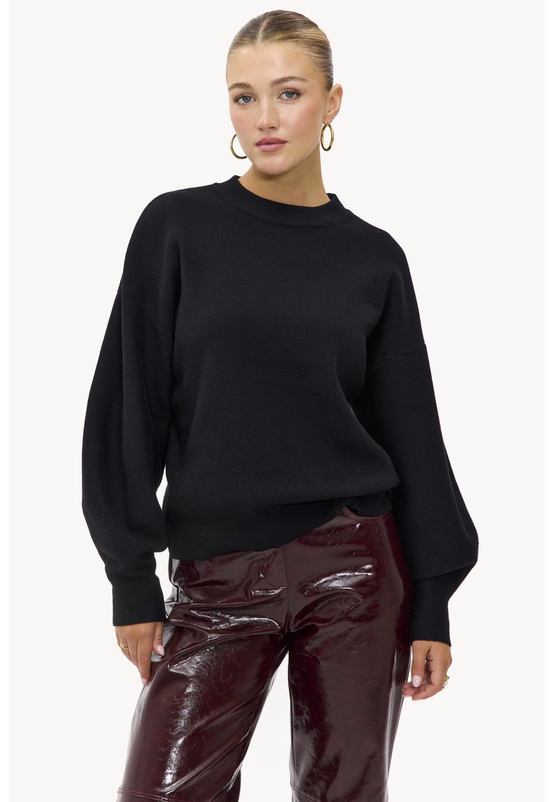 Pull noir en maille avec un col montant et des manches bouffantes, associé à un pantalon bordeaux brillant. Ourlet plus court à l'avant.