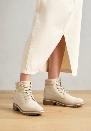 Béžové kotníkové boty vyrobené z texturovaného materiálu, s kulatou špičkou, šesti kovovými očky a robustní, vroubkovanou podrážkou.