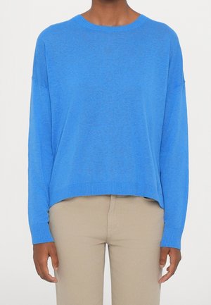 Personne portant un pull bleu vif à manches longues et un pantalon beige, debout devant un fond clair uni.