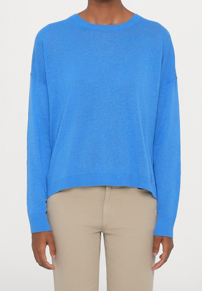 Personne portant un pull bleu vif à manches longues et un pantalon beige, debout devant un fond clair uni.