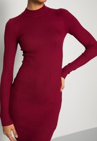Abito a coste bordeaux con collo alto e maniche lunghe, caratterizzato da una silhouette aderente e una texture sottile su tutta la superficie.