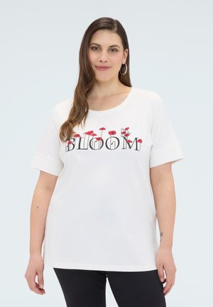 Kobieta z długimi brązowymi włosami nosi białą koszulkę z czerwonymi makami i napisem "BLOOM" na przodzie, czarne spodnie oraz kolczyki koła.