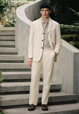 Young man standing on outdoor stairs wearing cream suit, checkered shirt, and brown shoes, surrounded by greenery and stone walls.