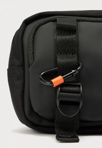 Μαύρη τσάντα με υφή ματ φινίρισμα· διαθέτει μαύρο καραμπίνερ με πορτοκαλί λεπτομέρειες και ιμάντα. Συμπαγής και ορθογώνιο σχήμα.