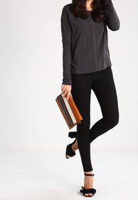 Maglia grigia a maniche lunghe con una linea centrale di borchie, jeans neri aderenti, tacchi neri con spalline a volant e una pochette a strisce marroni.
