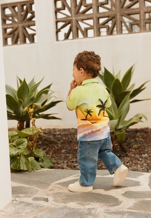 Lys grøn kortærmet polo skjorte med print af solnedgang og palmetræer, parret med blå jeans og hvide sneakers. Barnet går væk.