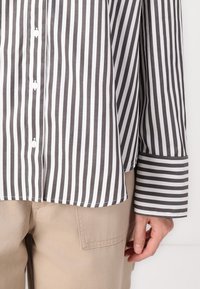 Striped button-up shirt featuring vertical black and white stripes, cotton material, rounded hem, and wide cuffs, paired with beige trousers.