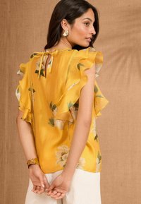 Blusa gialla con stampa floreale, maniche a ruota, dettaglio con laccetti sul retro e una texture liscia. Presenta motivi floreali bianchi e una vestibilità comoda.