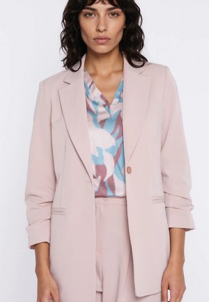Kobieta o ciemnych, falowanych włosach nosi jasnoróżowy blezer i pasujące spodnie na wzorzystej bluzce z abstrakcyjnymi kształtami w kolorach niebieskim, białym i mauve.