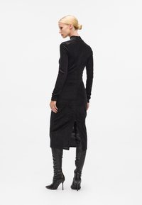 Vestido preto justo com mangas longas, tecido texturizado e uma fenda nas costas. Usado com botas pretas de salto alto até ao joelho.