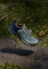 Chaussure de course verte et marine avec une tige en maille texturée, semelle rembourrée et accents marron, flottant au-dessus d'une surface en gravier.