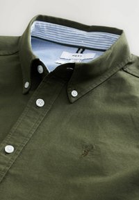 Chemise en coton vert olive avec un col boutonné, des boutons blancs et un col intérieur rayé bleu. Présente un petit motif brodé.