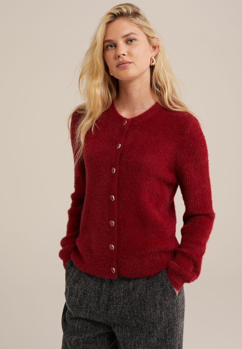 Sarkans pūkains kardigans ar noapaļotu apkakli un sešām metalliskām pogām priekšpusē. Redzama adīšanas tekstūra; piedurknes ir garas un nedaudz pieguļošas.