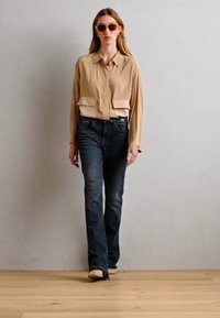 Beige skjorte med knapper og to brystlommer, kombinert med mørkeblå utsvingte jeans og lyse sko. Modellen har på solbriller.