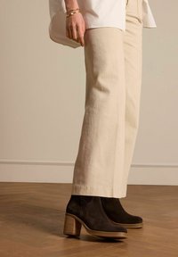 Zwarte suède enkellaarsjes met een robuuste houten hak, gecombineerd met crèmekleurige wijde broek, op een lichte houten vloer.
