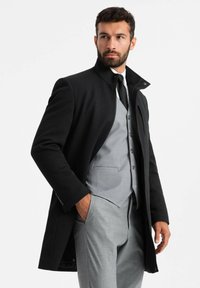 Cappotto nero con collo alto, stratificato su un gilet grigio chiaro e cravatta. Tessuto strutturato, vestibilità su misura, con chiusura a zip e tasche.