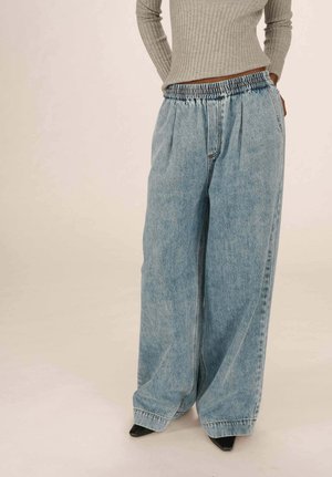 Personne portant un jean en denim bleu clair taille haute et ample avec une taille élastique, un pull beige côtelé, et des chaussures noires à talons pointus.